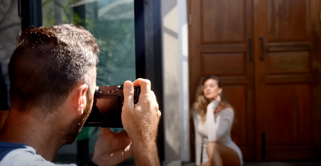 A man capturing a photograph of a girl sitting in front of a large door, with a white long-sleeve dress.