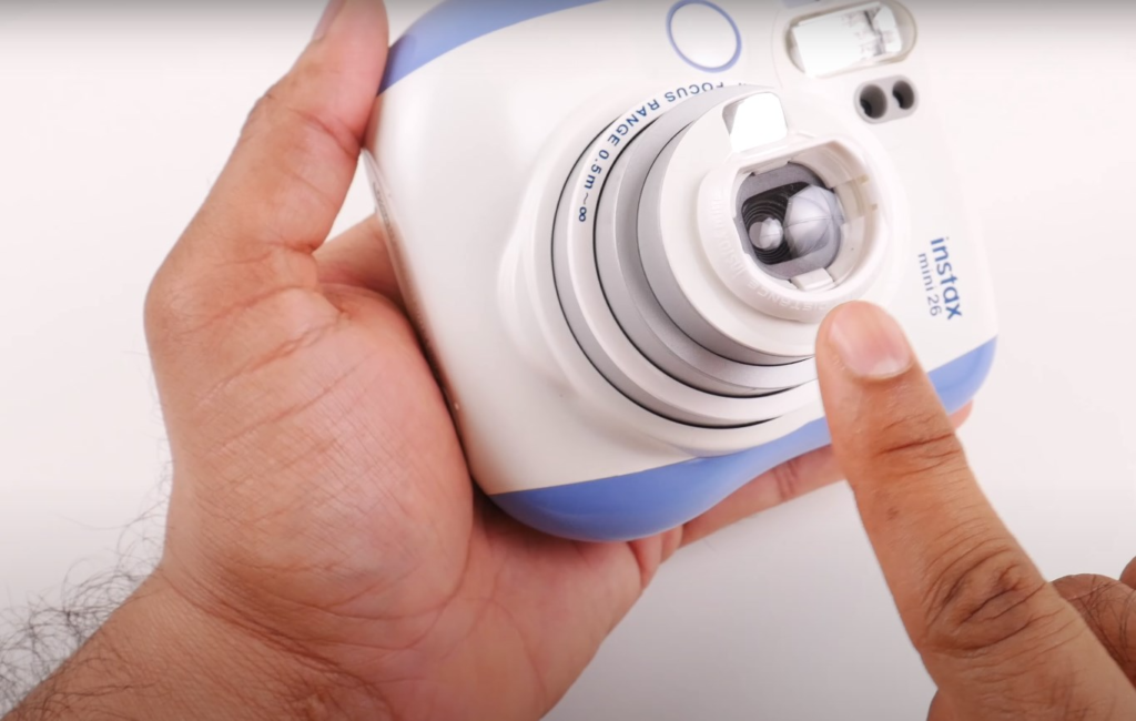 a hand holding the blue-and-white Instax 26 camera pointing at the lens