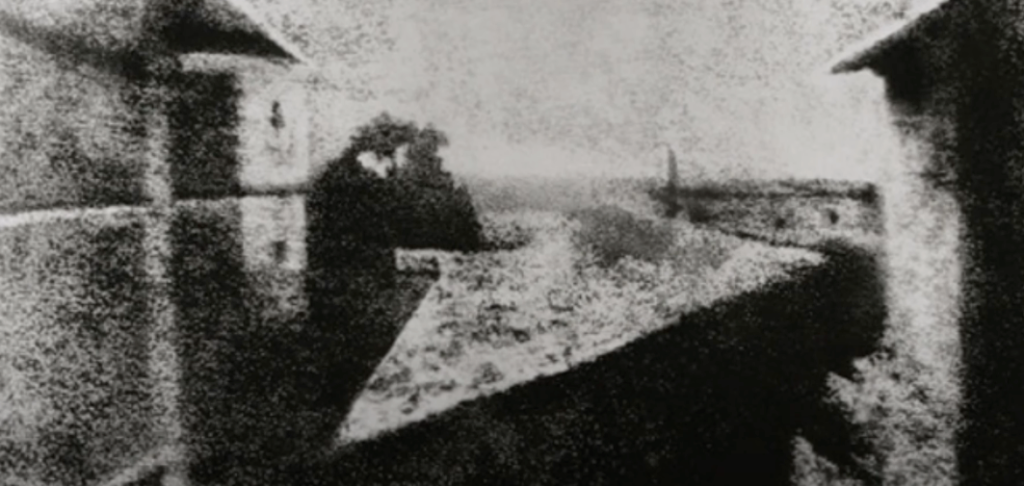 A blurry black and white screenshot of 'View from the Window at Le Gras,' capturing sections of buildings and the surrounding countryside.