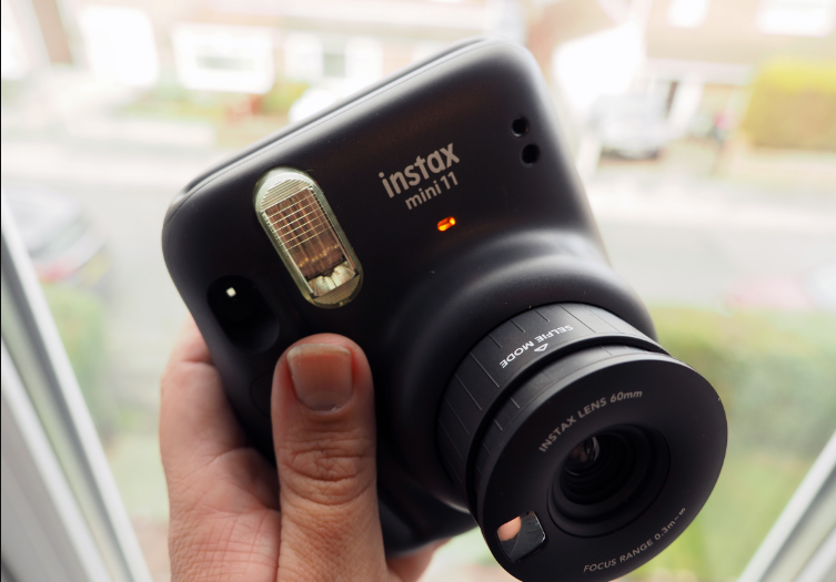 An Instax Mini 11 camera in a hand in front of a window