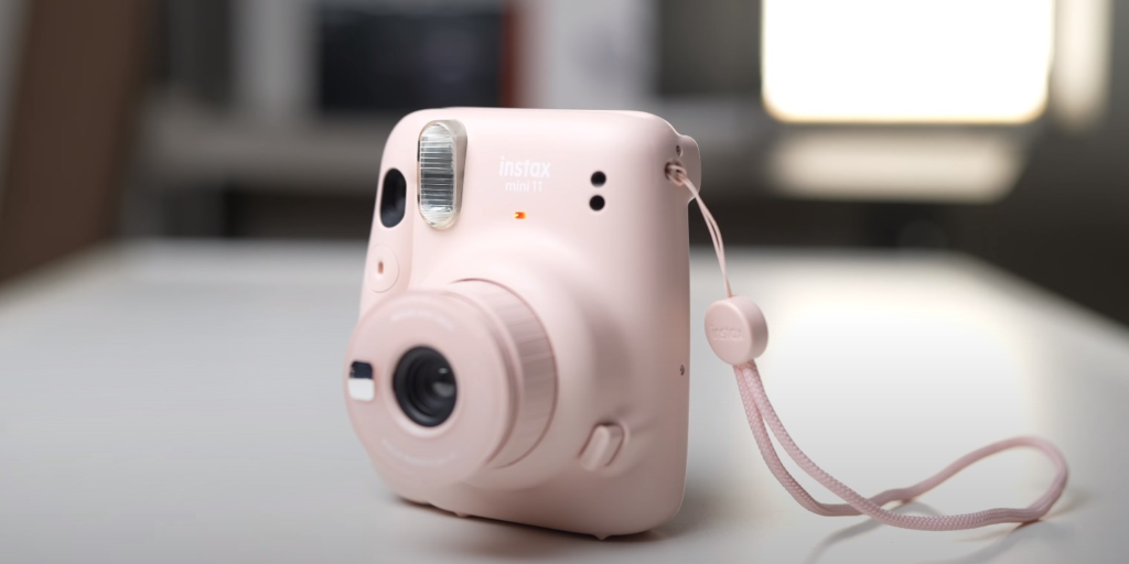 a pink Instax Mini 11 camera with a strap placed on a white table