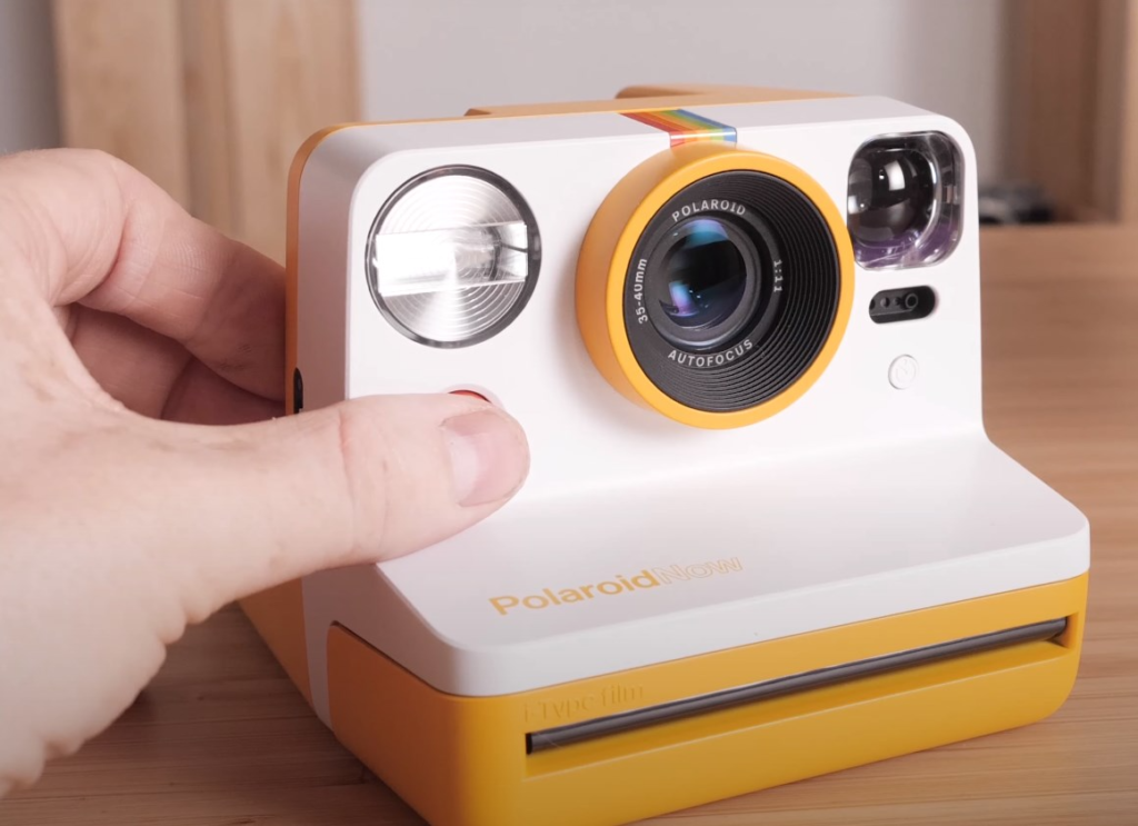 a hand pressing a button on the white/yellow Polaroid Now camera positioned at an angle