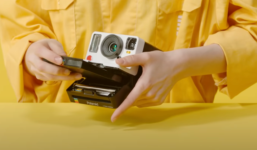 An image of a woman’s hand dressed in yellow loading a film into a Polaroid camera.