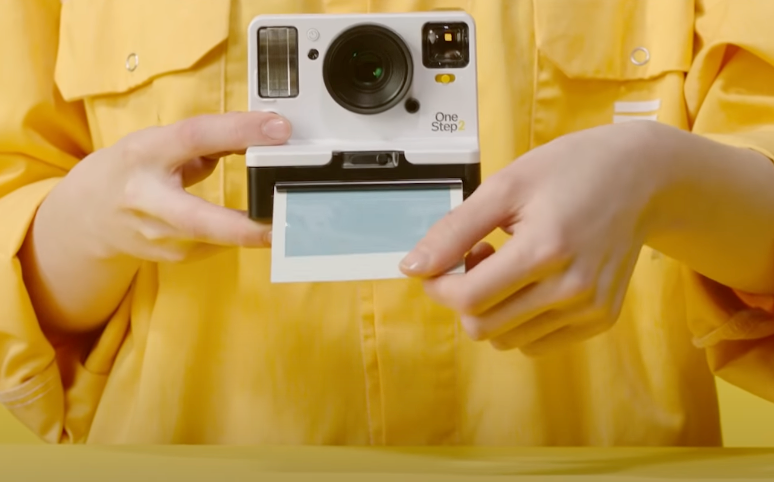 An image of a woman’s hand dressed in yellow developing a photo from a Polaroid camera.