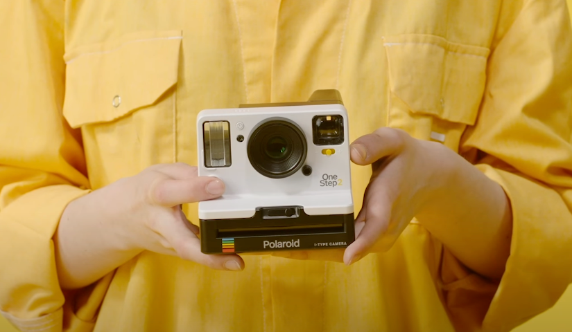 An image of a woman’s hand dressed in yellow holding a white Polaroid camera.